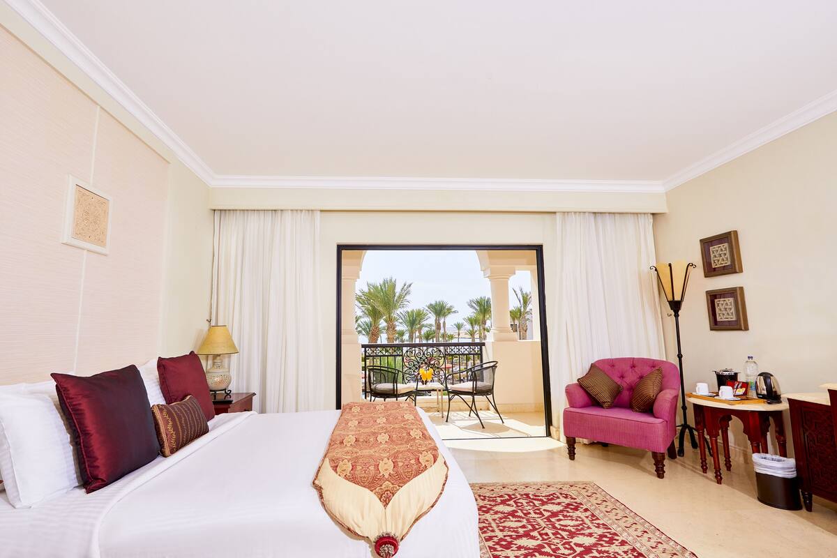 A hotel room with a large bed, a balcony with a view of palm trees, a pink armchair, and a small table with a lamp.