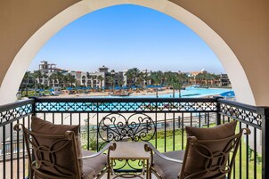 Un balcón con dos sillas y una mesa, con vista a una piscina y una playa con palmeras y edificios de un complejo turístico.