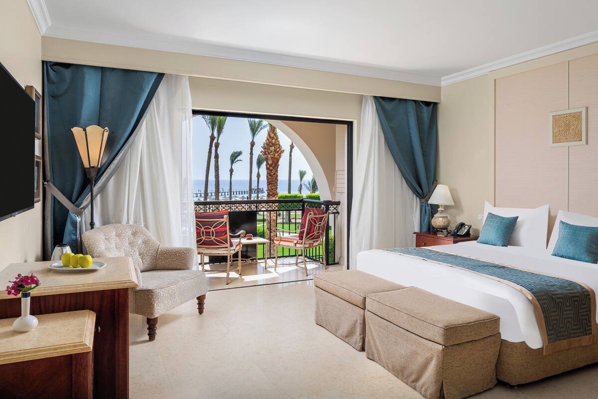 A hotel room with a large bed, a desk with a vase, a chair, and a view of palm trees and the sea through a balcony.