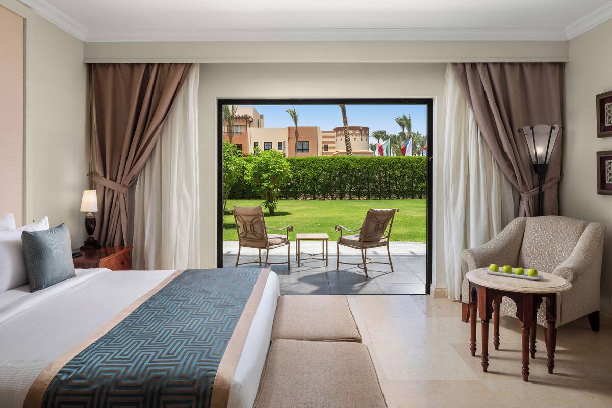 A hotel room with a large bed, a small table with fruit, two chairs, and a view of a garden and buildings outside.