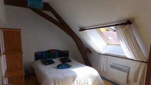 Een slaapkamer met een schuine plafond, een bed met witte beddengoed en kleurrijke kussens, een houten garderobe en een dakraam met gordijnen.