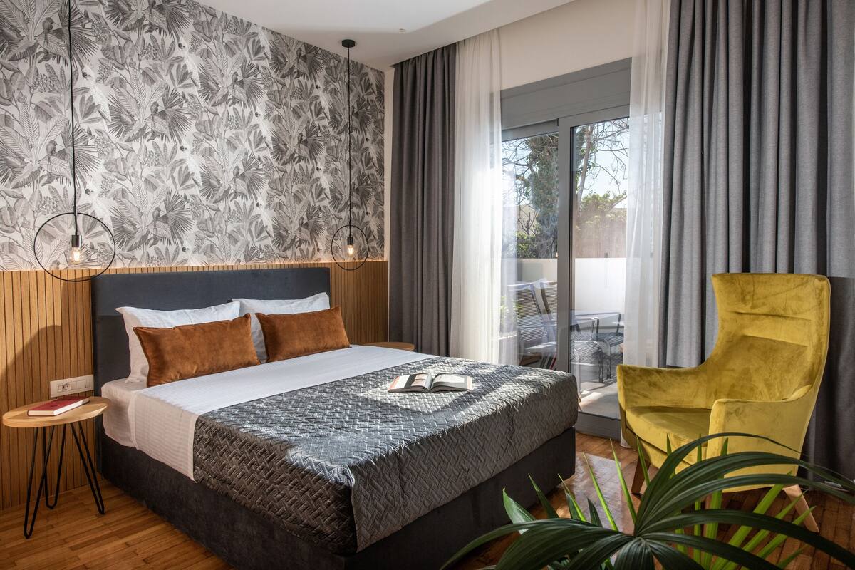 Een moderne hotelkamer met een groot bed, een houten hoofdeinde, een klein nachtkastje, een gele fauteuil en uitzicht op het omliggende landschap door een groot raam.