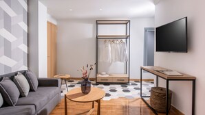 Een moderne woonkamer met een grijze bank, een ronde houten tafel met een vaas, een kledingrek, een televisie en een vloer met een patroon.