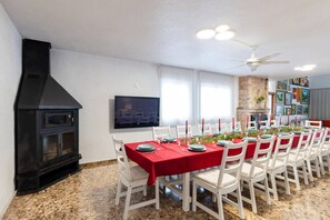 A dining area with a black wood stove, white chairs, a red tablecloth-covered table, and a television mounted on the wall.