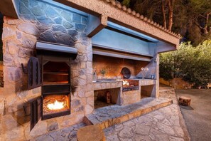 An outdoor stone fireplace with a fire, built into a stone wall with a blue awning above. - Fidalsa Paradise Pines (La Alcayna)