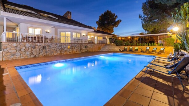 A swimming pool with a stone wall and a tiled deck, surrounded by lounge chairs and a building with a tiled roof. - Fidalsa Paradise Pines (La Alcayna)