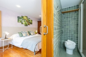 Habitación de hotel con cama, mesita de noche con lámpara, baño con inodoro y ducha, y zona de ducha con azulejos verdes.