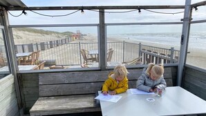 Two children at a wooden table with papers, overlooking a beach with a wooden walkway.