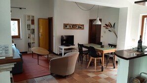Un soggiorno con TV, tavolo da pranzo e sedie. Si nota una libreria e una porta che dà su un'altra stanza.