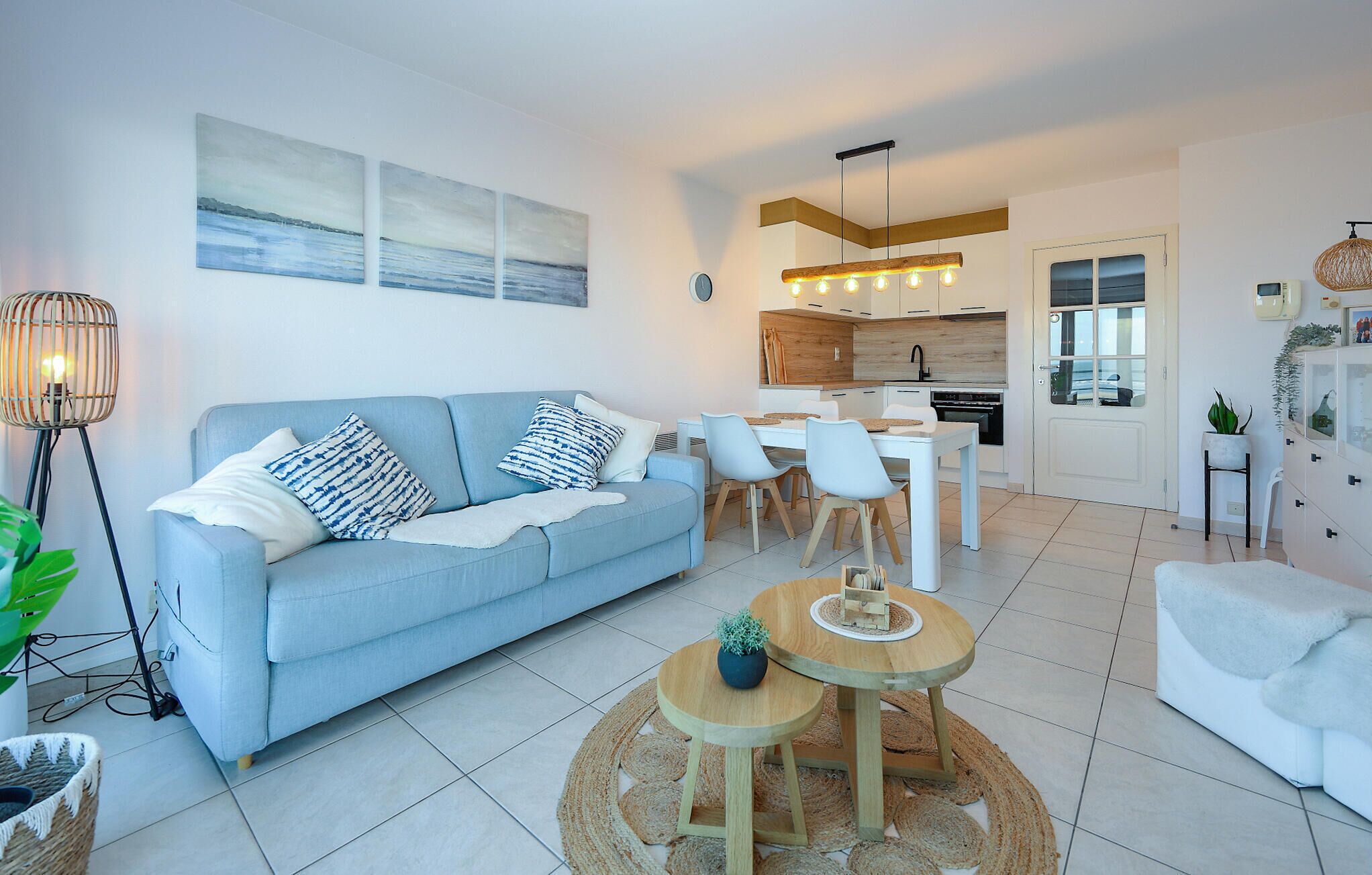 A modern living room with a light blue sofa, a round wooden coffee table, and a dining area with white chairs.