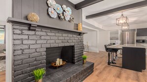 A modern living room with a black brick fireplace, a dining area with a large island, and a kitchen with stainless steel appliances. - 4BR w heated pool, basketball hoop, patio (Mesa)