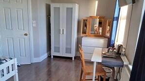 Une pièce de taille modeste, dotée d’un plancher en bois, d’un bureau blanc avec une chaise, d’une armoire, d’un miroir et d’un téléviseur fixé au mur.