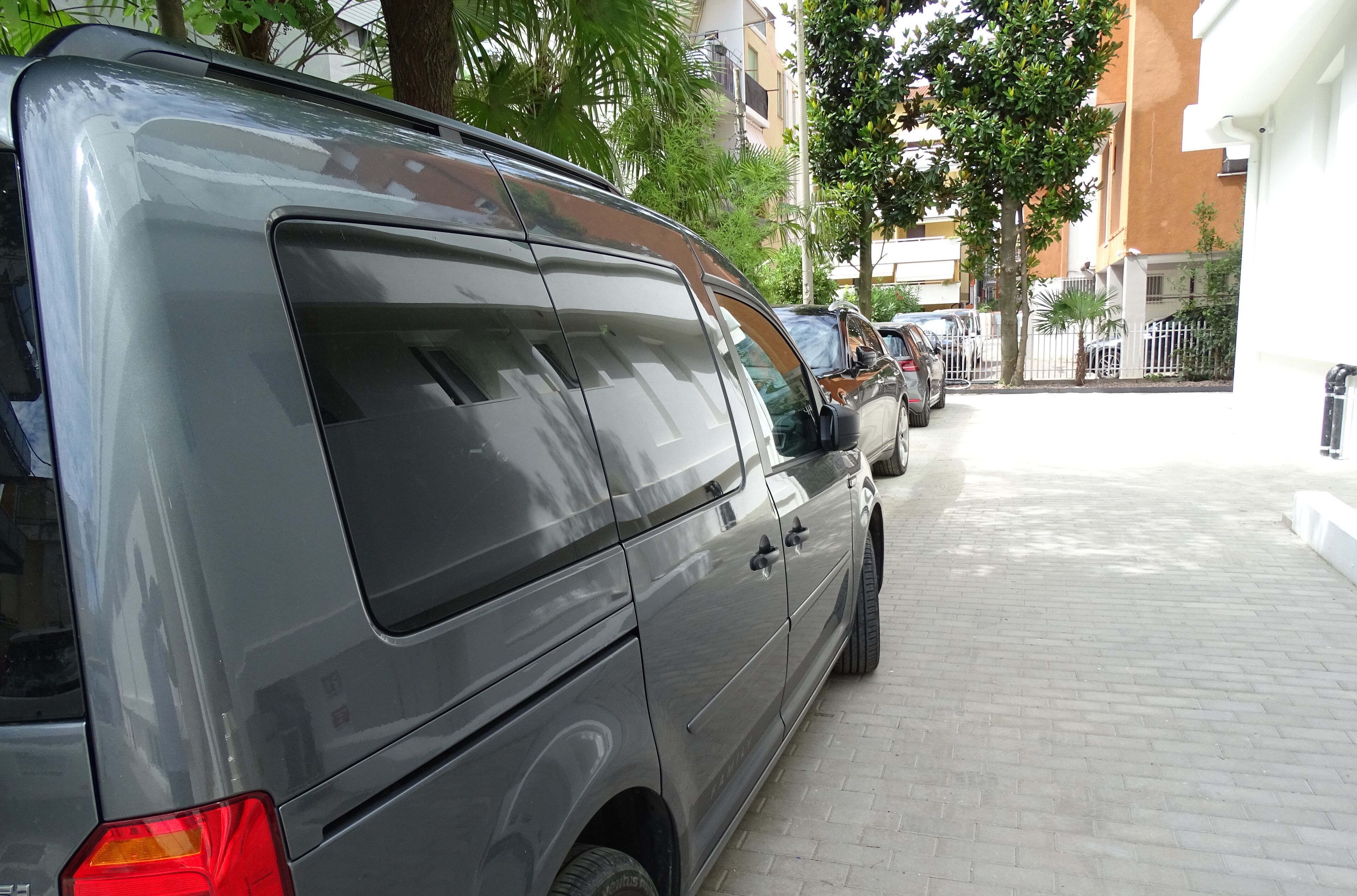 A grey van parked on a paved sidewalk next to a building with a white exterior.