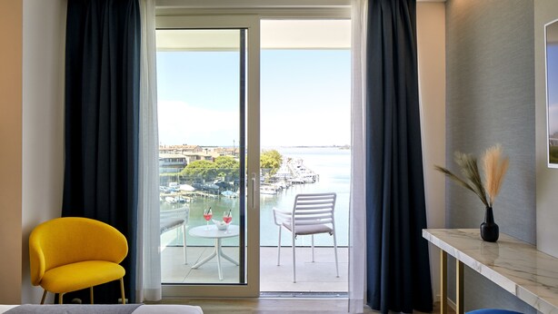 Habitación de hotel con balcón, una cama, una silla amarilla, una silla blanca y una pequeña mesa con dos copas de vino tinto.
