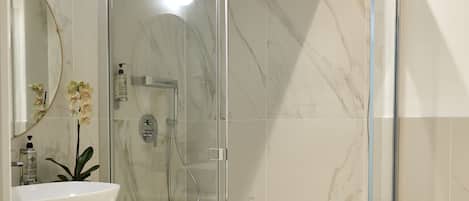 Un baño moderno con ducha envuelta en vidrio, piso de mármol y un lavamanos con espejo superior.