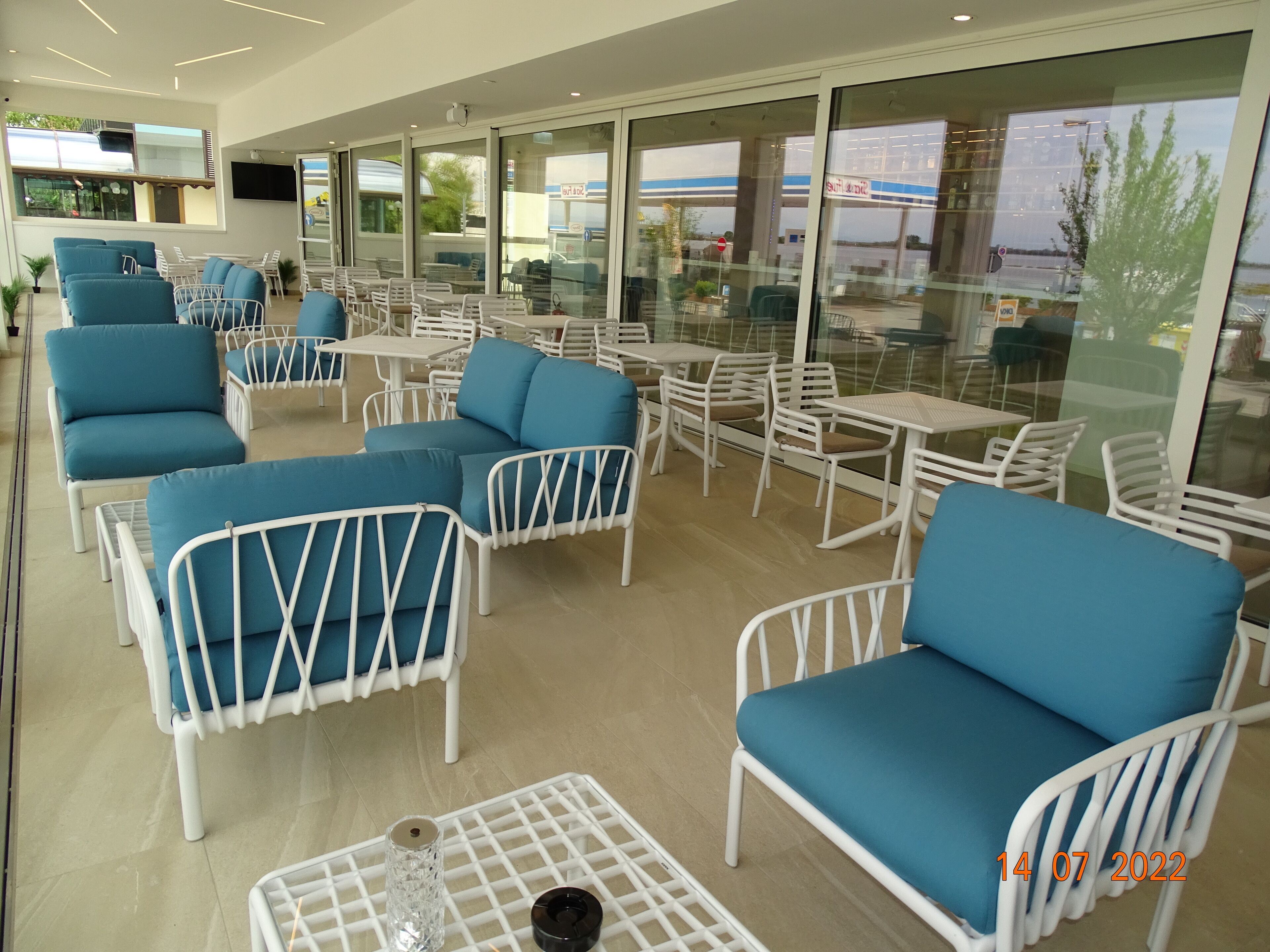 A modern outdoor seating area with blue and white chairs, tables, and a glass wall offering views of the outside.