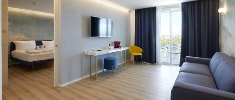 A modern hotel room with a bed, a desk, a flat-screen TV, a sofa, and a balcony with curtains.