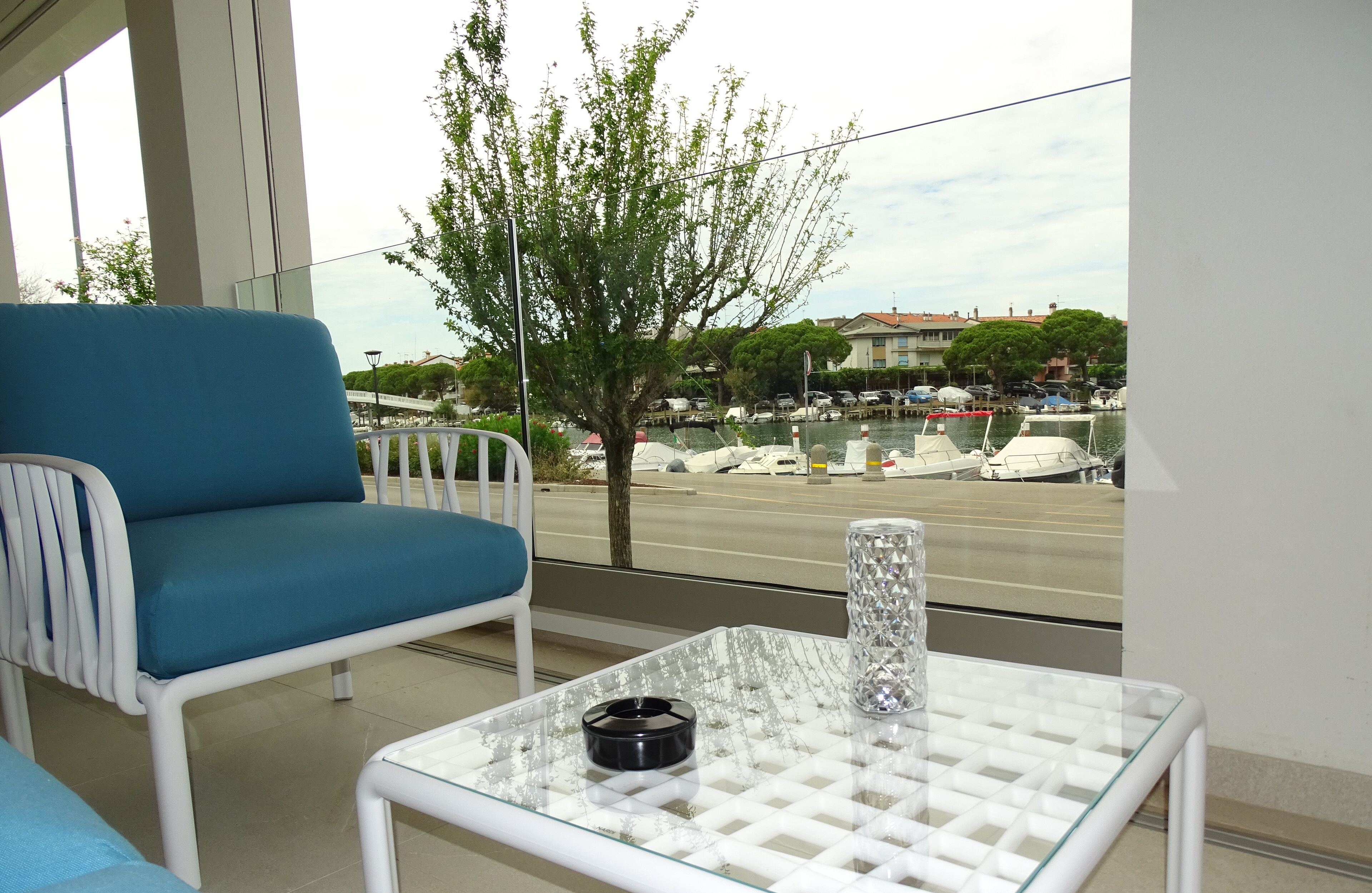A balcony with a blue chair, a glass table with a cigarette, and a view of a marina with boats.