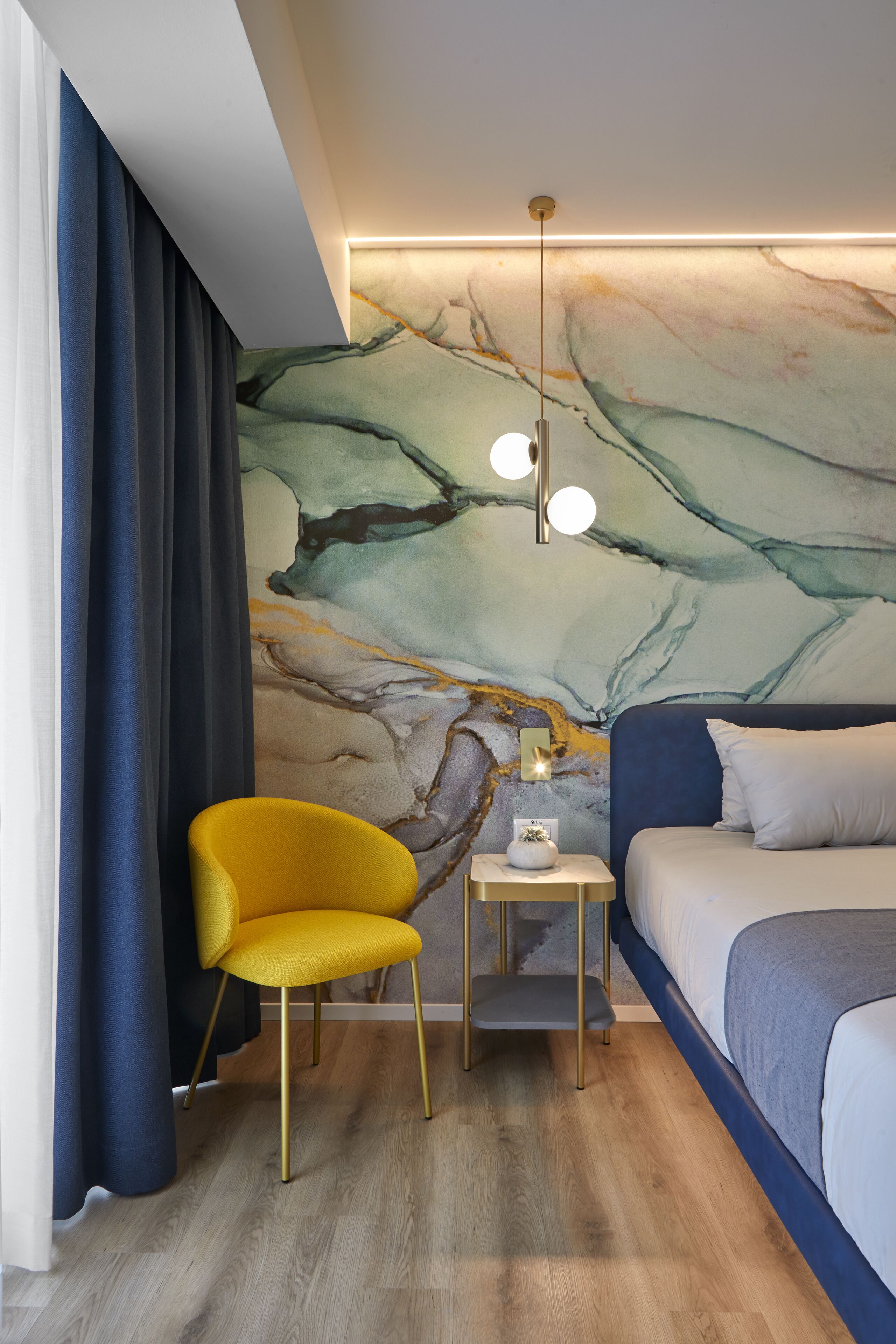 Une chambre d’hôtel moderne avec un mur décoré d’un motif marbré, une chaise jaune, une table de chevet avec une lampe et un lit avec une literie bleue.