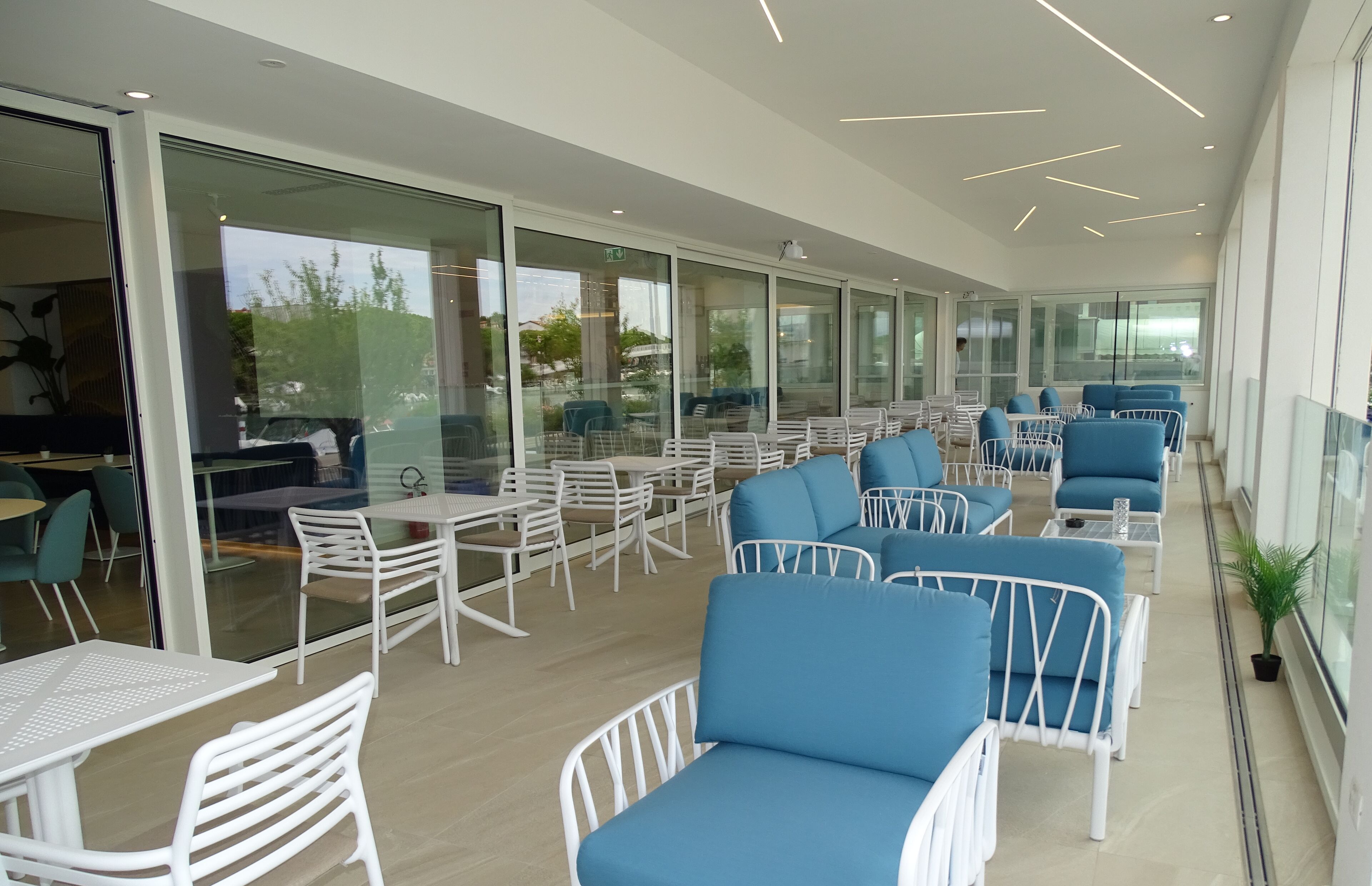 A modern outdoor seating area with white metal chairs and blue cushioned sofas, large glass windows, and a clean, contemporary design.