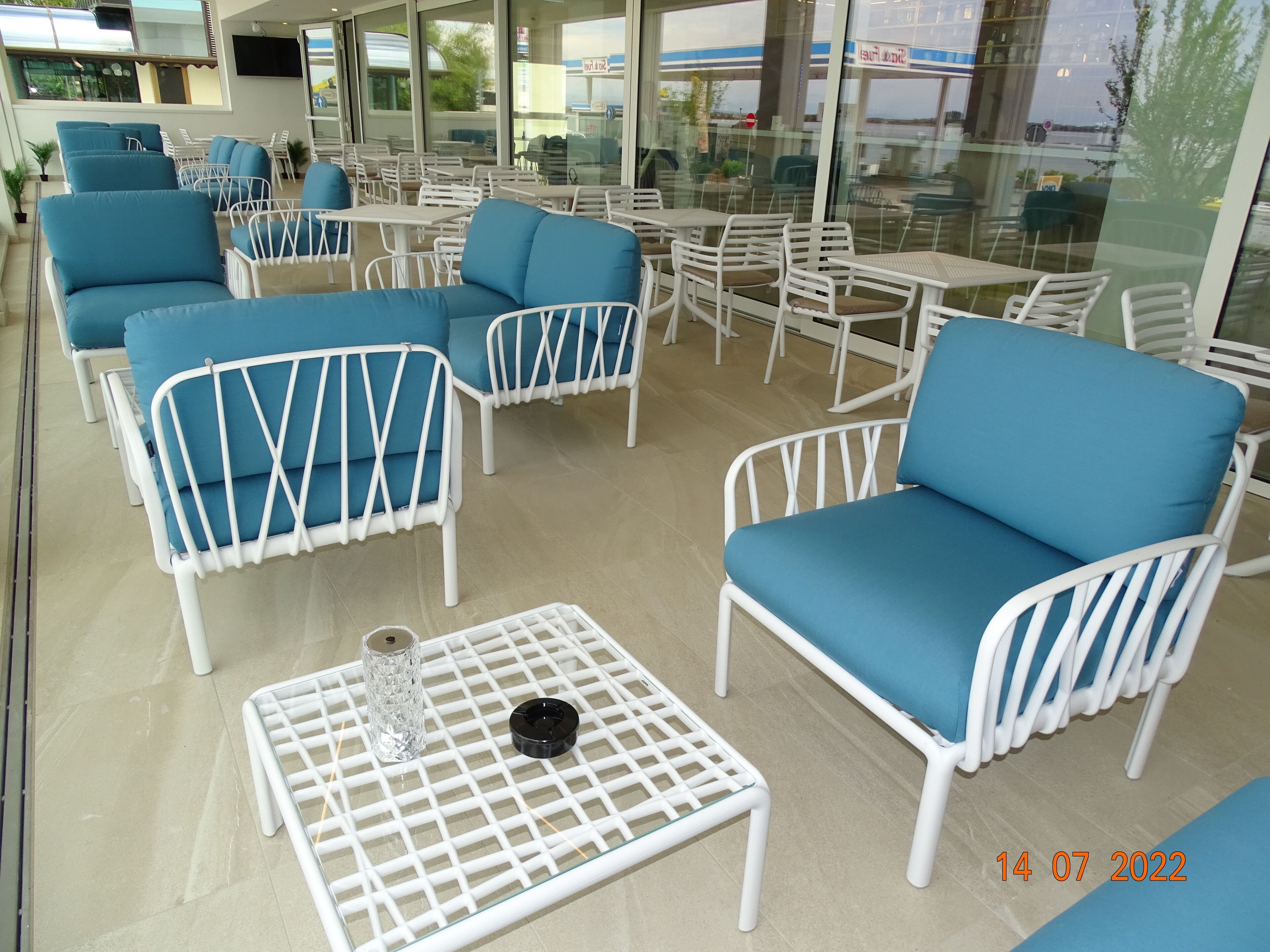 A seating area with white metal chairs and blue cushioned sofas, a glass coffee table, and a view of the outside.