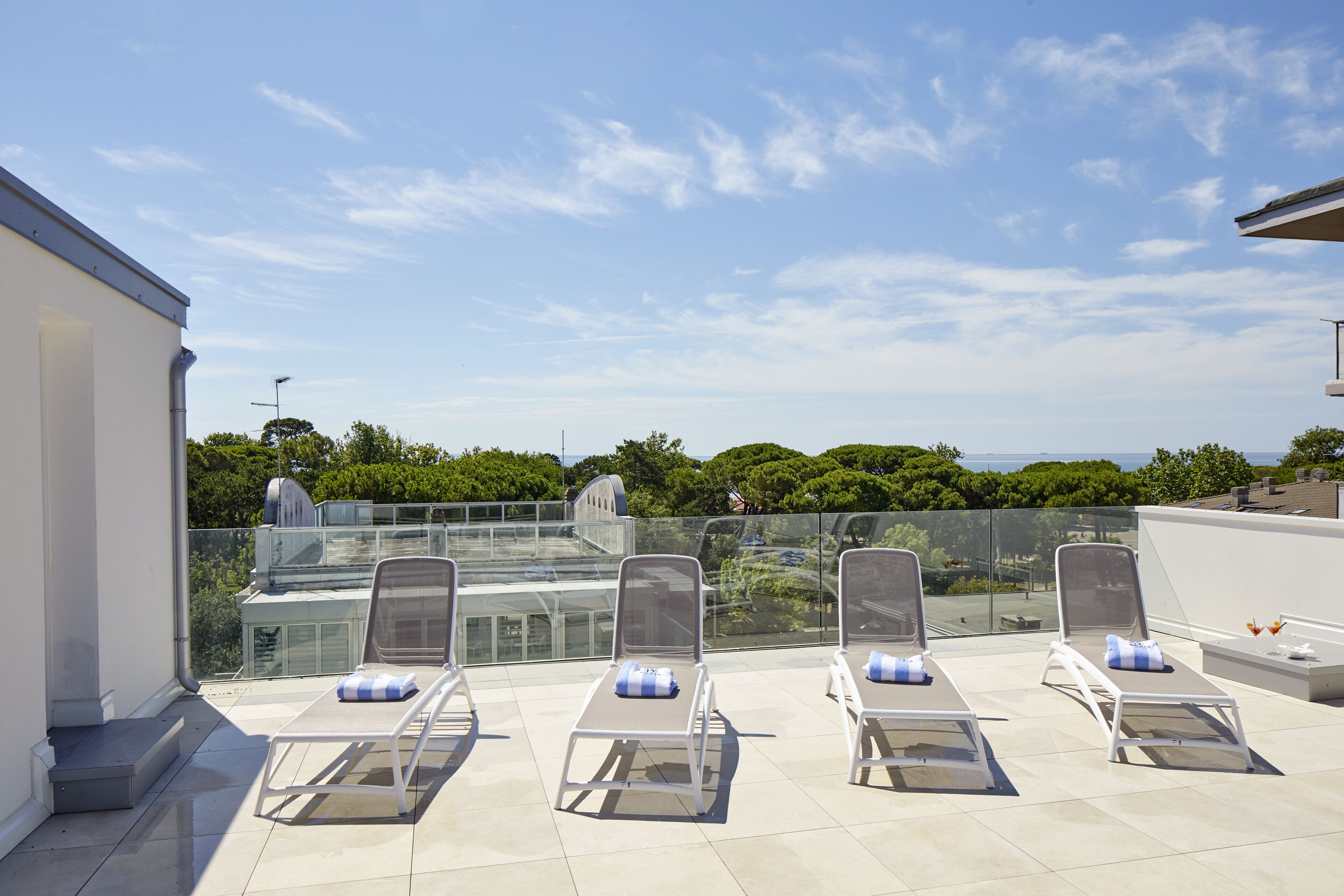 Terraza en la azotea con cuatro tumbonas, barandilla de cristal y vistas al mar y vegetación.