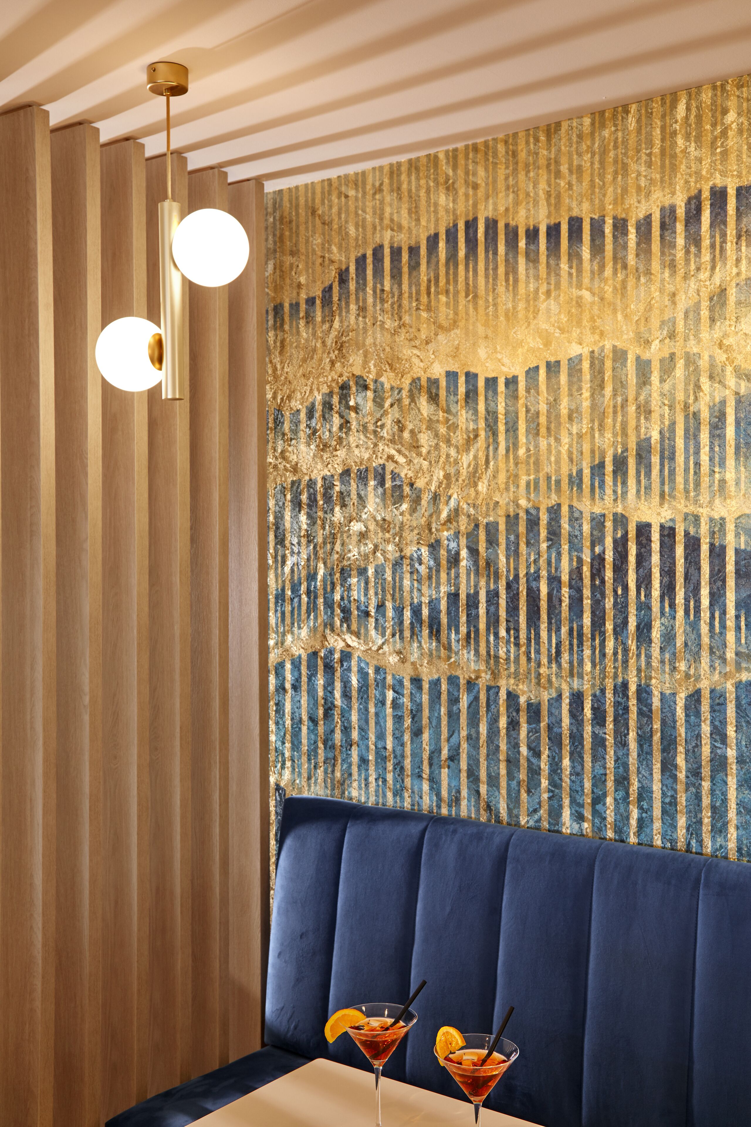A restaurant interior with a blue upholstered booth, two cocktails on the table, and a decorative wall with vertical stripes.