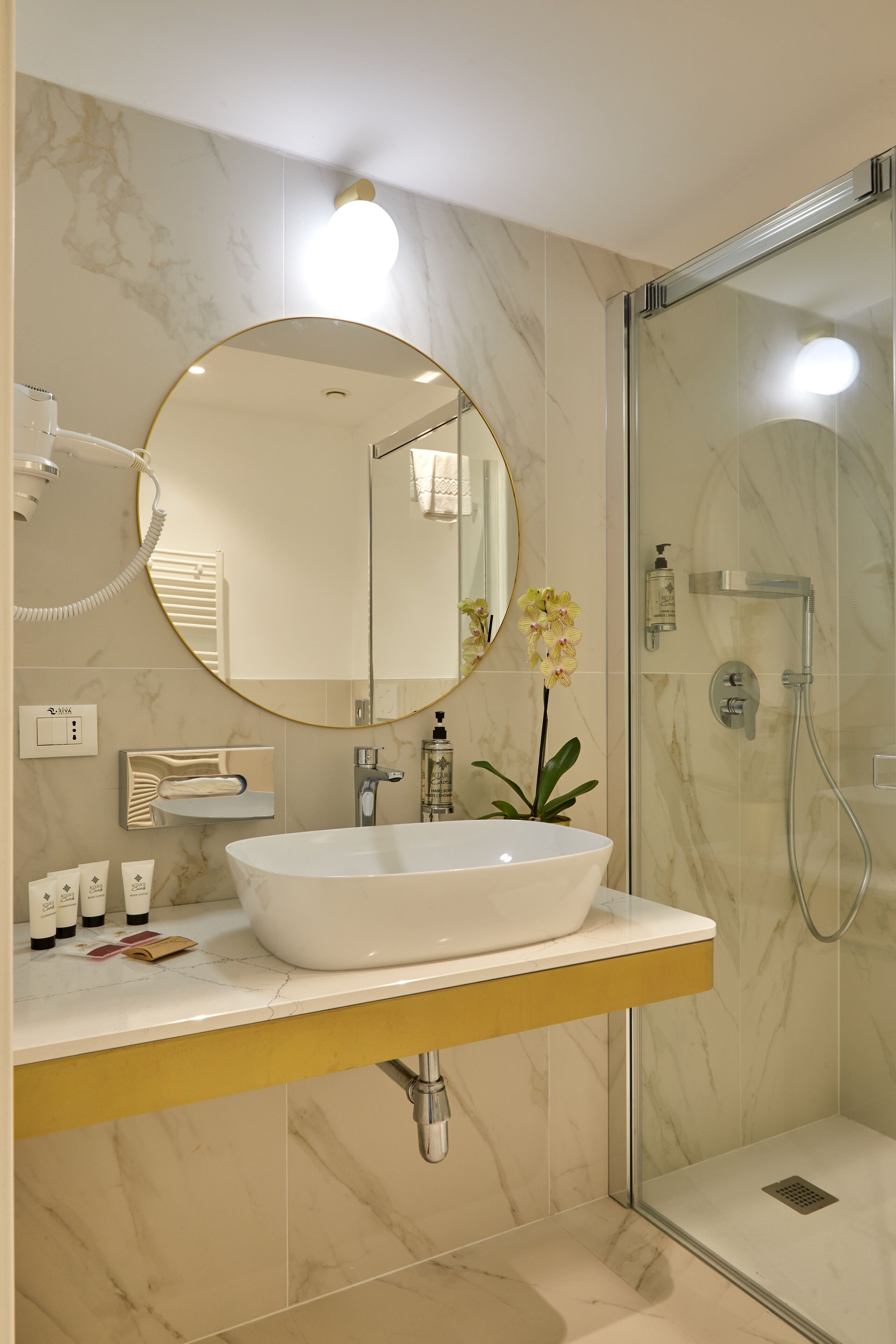 A bathroom with a white sink, a round mirror, a shower, and a yellow plant.