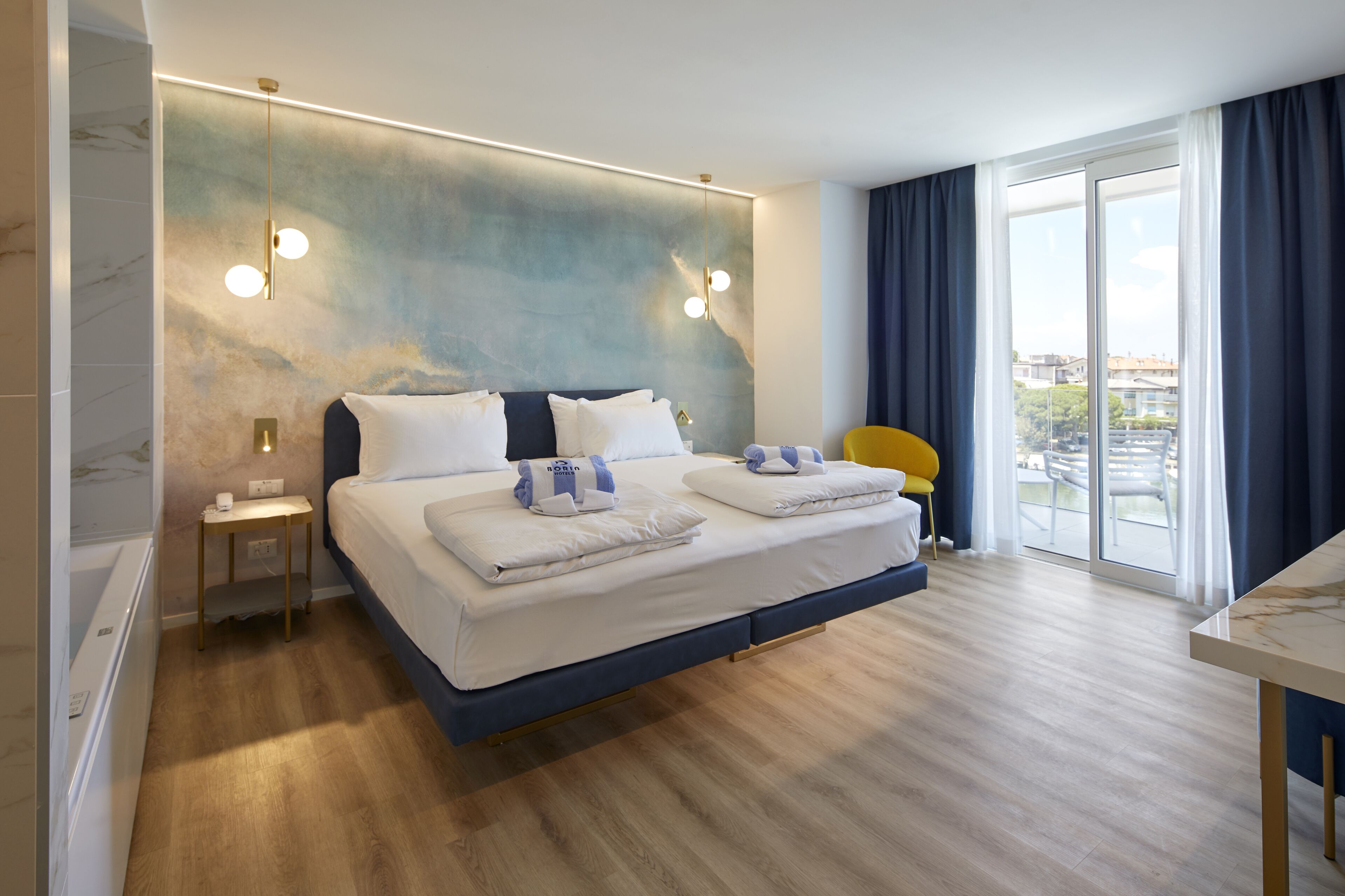 A modern hotel room with a large bed, a small table, a chair, and a view of the city through the window.