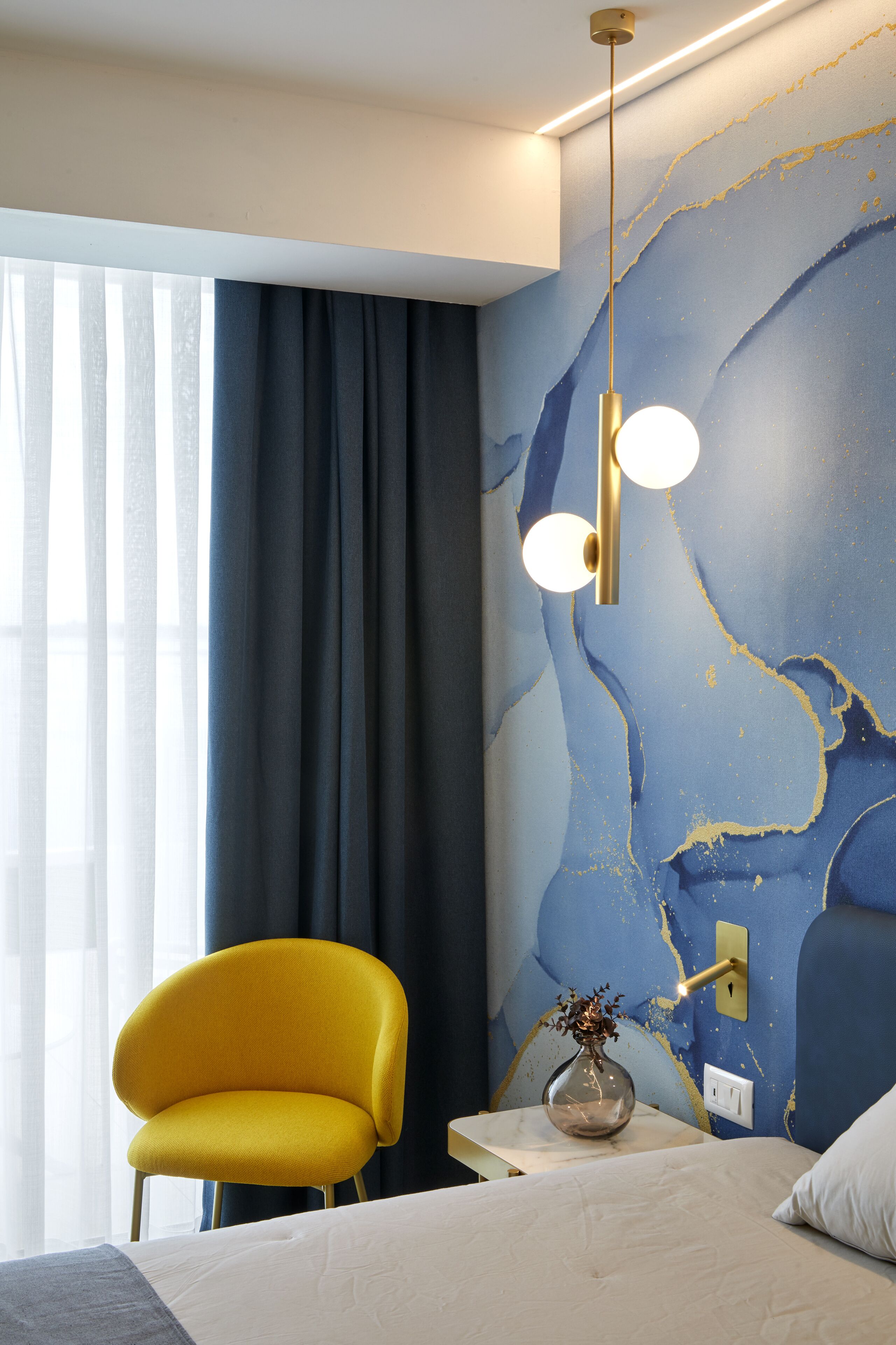 A modern hotel room with a bed, a yellow armchair, a vase with flowers, and a wall with a blue and gold abstract design.