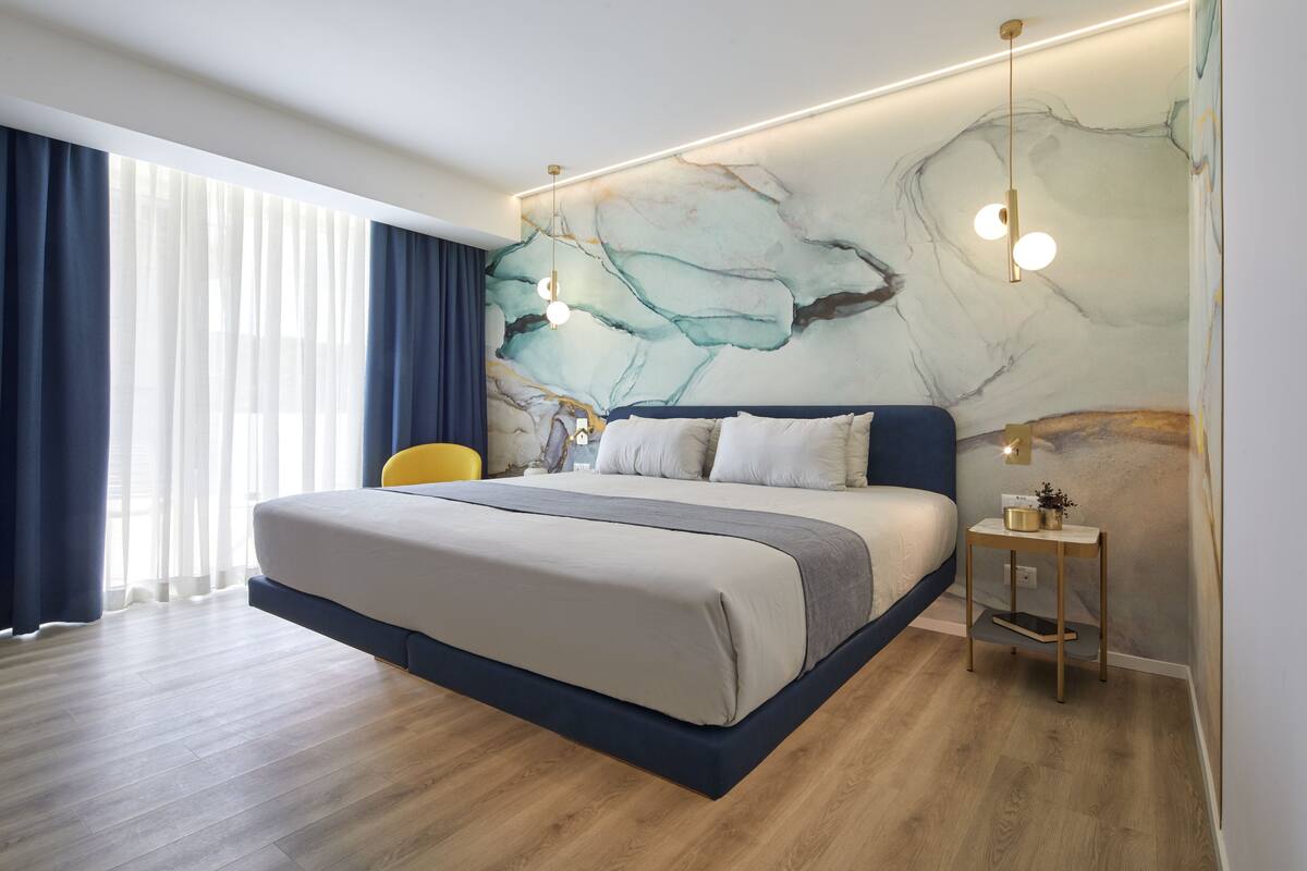 A modern hotel room with a large bed, a nightstand, a wall-mounted light fixture, and a view of the outdoors through sheer curtains.