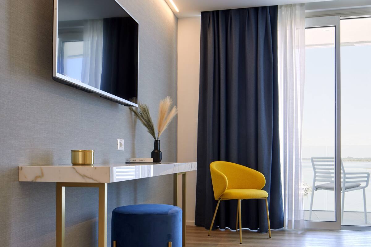 A modern hotel room with a flat-screen TV mounted on the wall, a small table with a vase, a blue ottoman, a yellow chair, and a large window with curtains.
