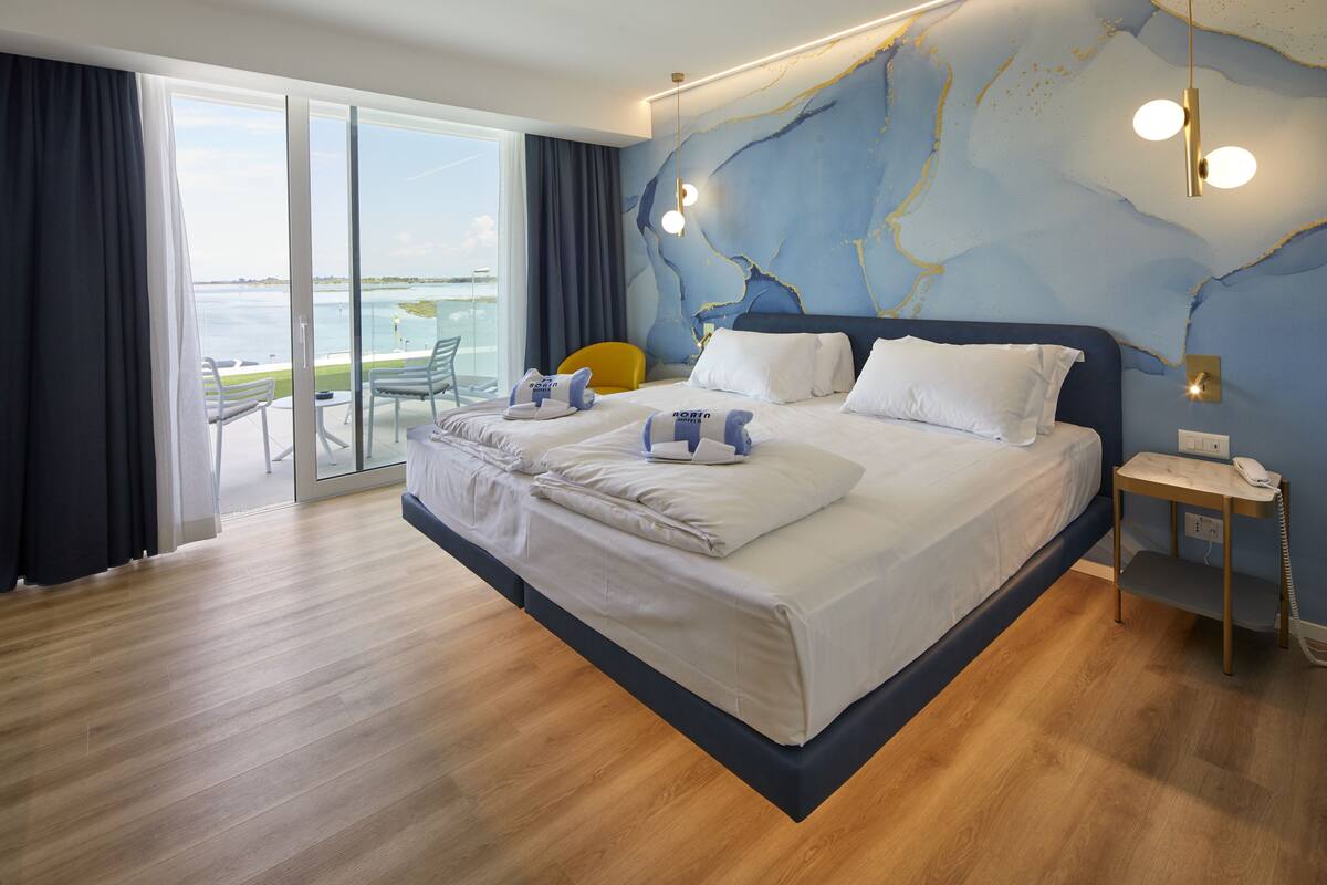 A modern hotel room with a large bed, a view of the sea through a sliding glass door, and a wooden floor.