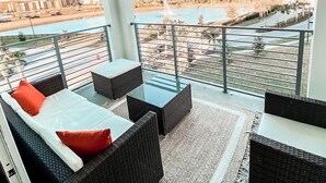 Un balcón con mobiliario de mimbre, una mesa de vidrio y vista a un río y edificios.