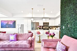 A modern living room with a pink sofa, a coffee table, and a TV. There is a kitchen area with white cabinets and a green wall. - The Princess Penthouse with lazy river and close to Disney World (Kissimmee)