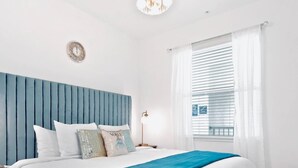 Un dormitorio con una cama, cabecera azul, candelabro, reloj y ventana con persianas.