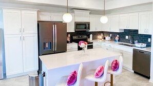 Una cocina moderna con armarios blancos, un refrigerador de acero inoxidable, un microondas y un lavamanos. Hay tres taburetes de bar con cojines rosados.