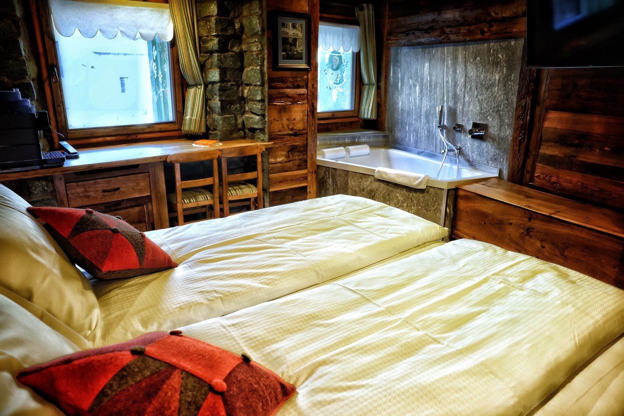 Une chambre confortable, aménagée dans le style d’un chalet, avec un grand lit, un bureau en bois, un mur en pierre et une fenêtre avec des rideaux.