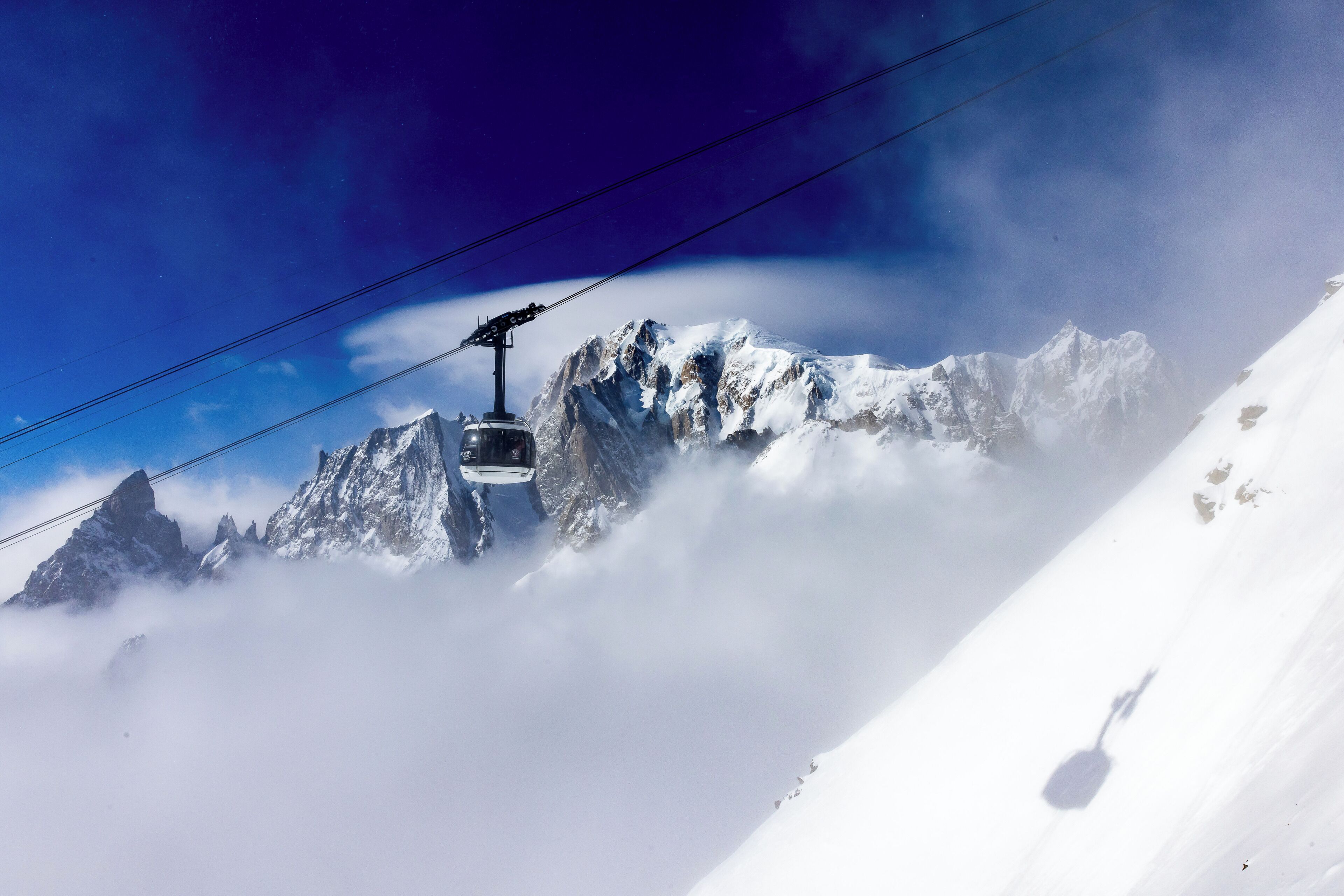 Un téléphérique qui monte une montagne enneigée aux pics acérés, avec des nuages en contrebas.