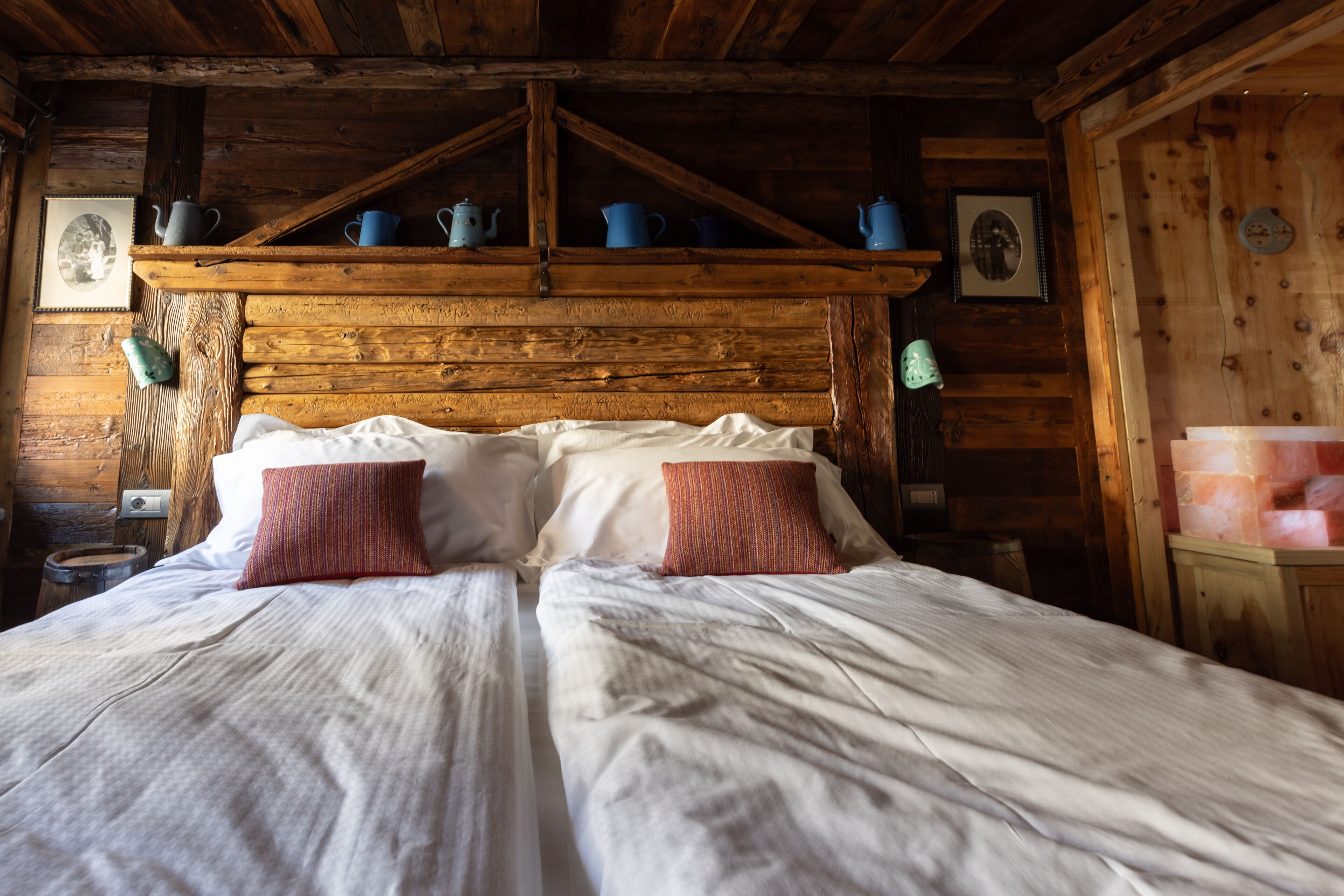 Une chambre dans une cabane en bois, avec un lit, des oreillers et une étagère où sont posés des pots.