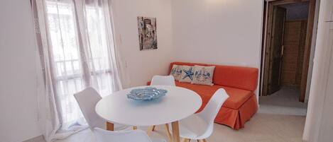 A dining area with a round table, white chairs, and a blue bowl. There's a red sofa with decorative pillows and a wall-mounted picture.