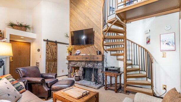 A living room with a fireplace, leather chairs, a wooden coffee table, a TV, and a spiral staircase. - West Keystone Condo w/ Quiet Location and Wrap Around Porch, Mins to Shuttle! (Keystone)