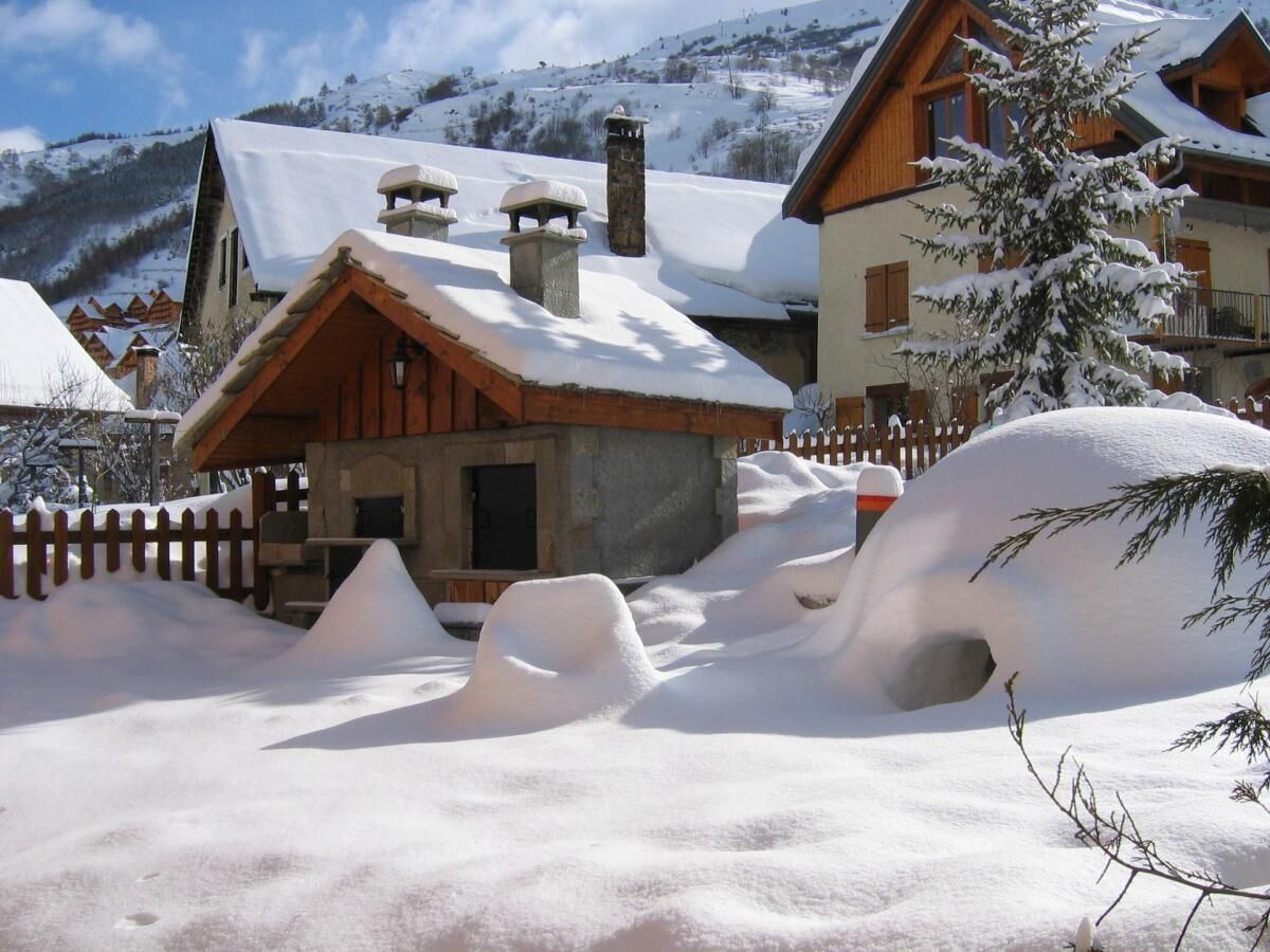A chalet-style building with a sloped roof and stone chimneys, surrounded by a wooden fence and snow-covered ground.