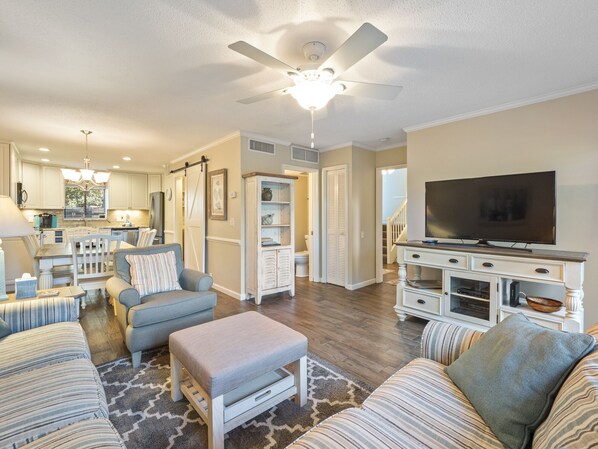 A spacious living room with a large TV, comfortable seating, and a kitchen in the background.