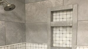 Une salle de bain carrelée de carreaux gris, une douche avec un pommeau de douche amovible et une petite fenêtre à motifs géométriques.