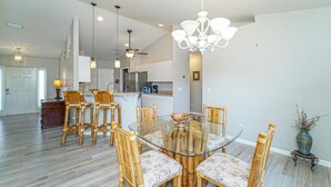 A spacious dining area with a glass table surrounded by wooden chairs, a chandelier, and a kitchen in the background. - Calling all boaters and fishermen!  Canal front home with 10k lb boat lift! (North Fort Myers)
