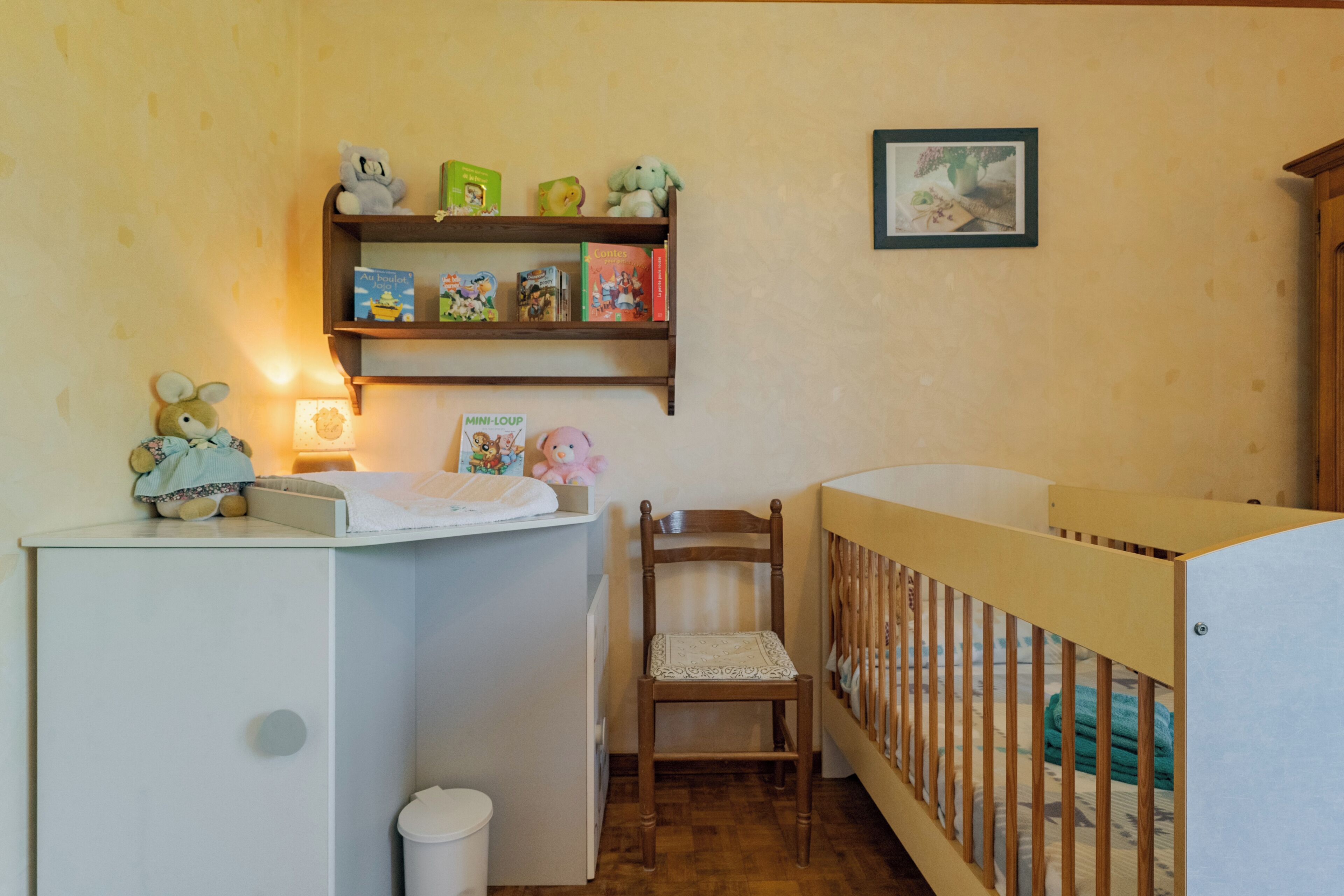 A baby room with a crib, a changing table, a shelf with books and toys, and a chair.