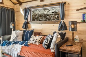 Interior acogedor de una cabaña con detalles de madera, una cama con muchos almohadones, una ventana con cortinas y dos lámparas fijadas a la pared.