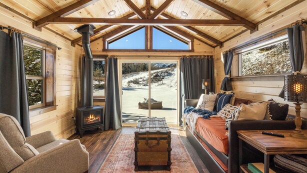 Interior acogedor de una cabaña con chimenea, un sofá, un cofre de madera y vistas a un paisaje nevado a través de amplios ventanales.