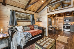 Ein gemütliches Blockhaus-Interieur mit Holzvertäfelung, einer Couch mit verschiedenen Kissen, einem Esstisch und einer Küchenzeile mit Herd und Backofen.